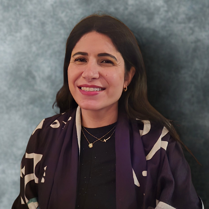 Portrait of a woman with long dark hair smiling warmly. She is wearing a dark outfit with layered gold necklaces and a patterned shawl, standing in front of a soft gray background.